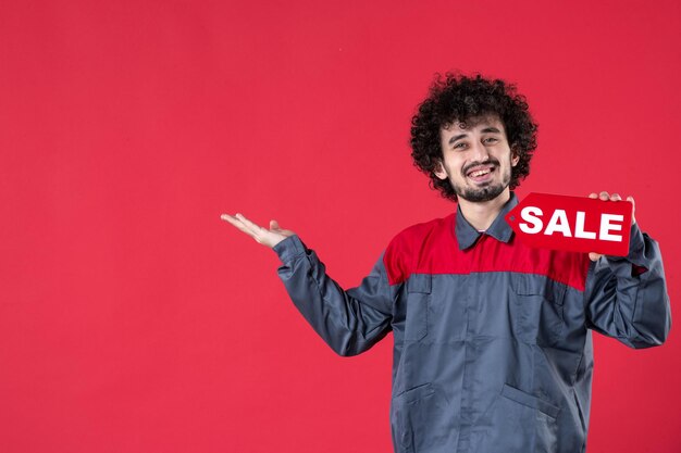 Front view male worker holding red sale nameplate on red background mechanic photo house uniform instrument color job worker