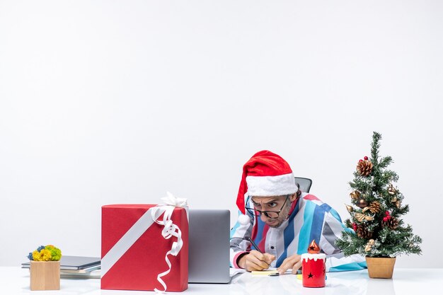 Вид спереди мужчина-работник, сидящий на своем месте с ноутбуком и файлами, пишущий заметки, офисная праздничная работа, рождество