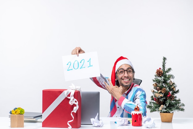 Front view male worker sitting in his working place happily holding paper sheet with number 2021, new year concept office job xmas