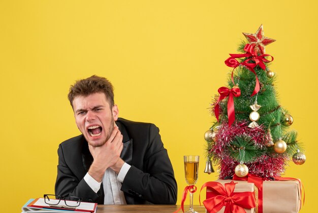 Front view male worker sitting behind his working place having throat pain on yellow 