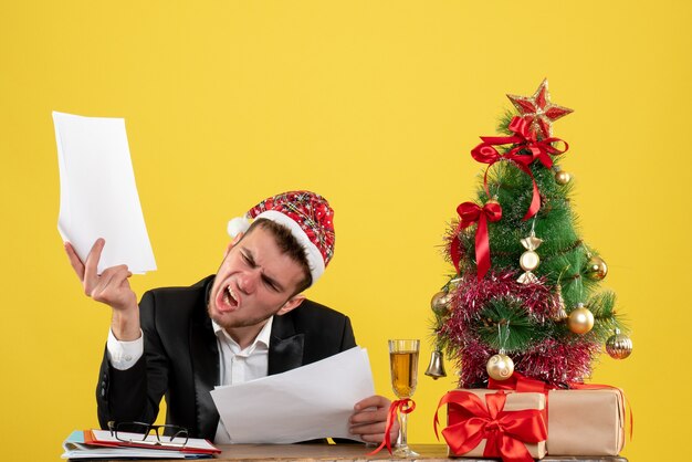 Front view male worker sitting behind his working place holding documents on yellow floor new year office color job christmas