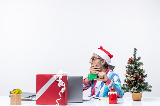 Вид спереди мужчина-работник, сидящий на своем рабочем месте, держа зеленую банковскую карту в офисе, рождественские деньги, работа, эмоция