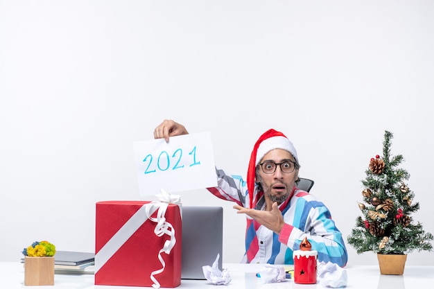 Free photo front view male worker sitting in his working place holding paper sheet with number 2021, new year concept