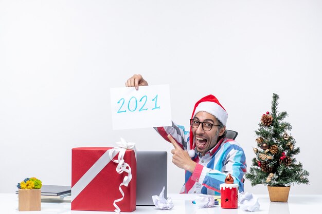 Front view male worker sitting in his working place holding paper sheet with number 2021, new year concept