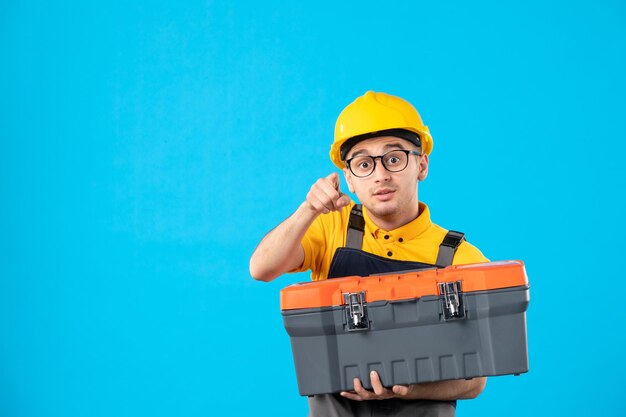 Foto gratuita vista frontale del lavoratore di sesso maschile in uniforme e casco con cassetta degli attrezzi nelle sue mani sull'azzurro