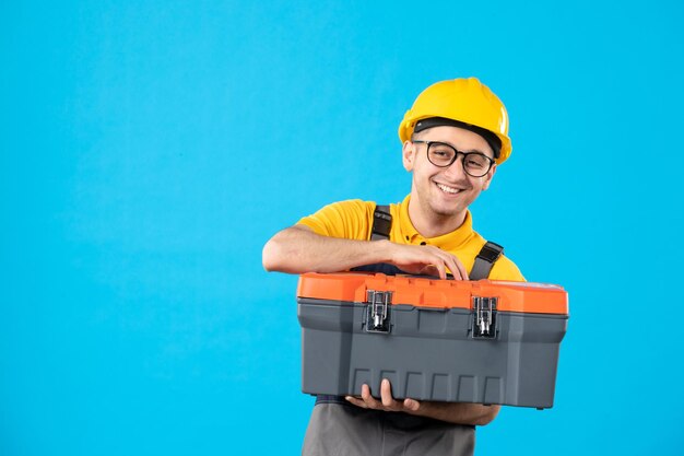 Front view of male worker in uniform and helmet with tool box in his hands on blue 