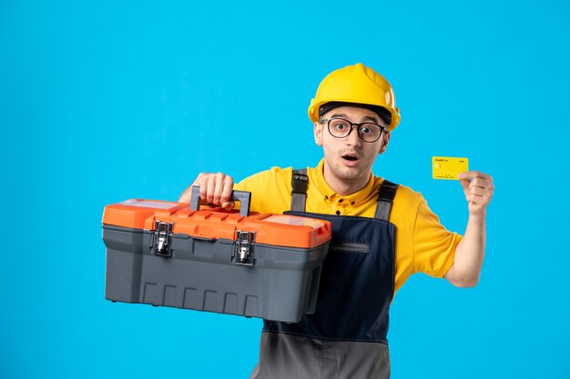 Front view male worker in yellow uniform with tool box and credit card on blue 