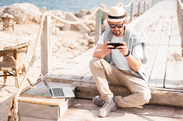 ノートパソコンとスマートフォンに取り組んでいるビーチの桟橋の男の正面図