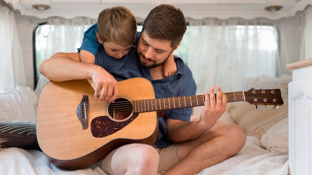 Front view man playing the guitar in a caravan next to his son