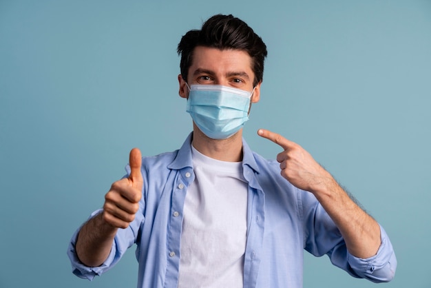 Front view of man pointing at the medical mask he's wearing and showing thumbs up