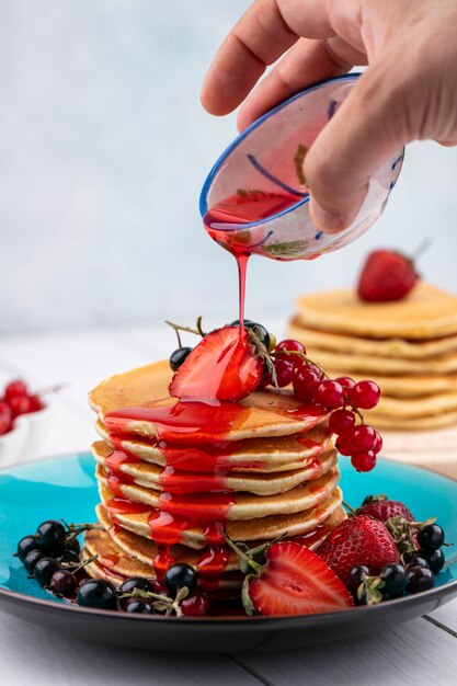 Front view a man pours strawberry syrup pancakes with strawberries black and red currants on a plate