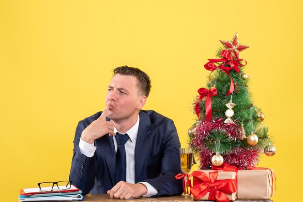 Front view of man in suit putting finger to his mouth sitting at the table near xmas tree and gifts on yellow