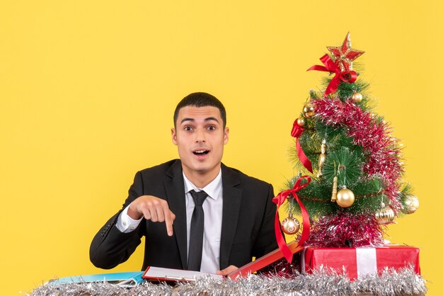 指のドキュメントのクリスマスツリーとギフトを指してテーブルに座っているスーツの正面図の男