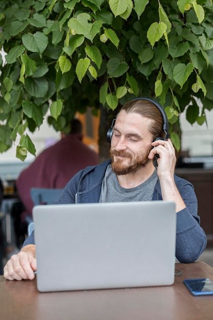 Vista frontale dell'uomo ad un terrazzo che ascolta la musica sulle cuffie con il computer portatile