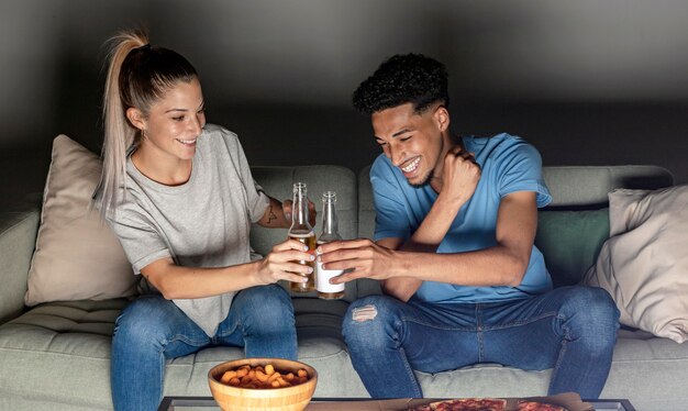 Front view of man and woman toasting with beer at home while watching tv