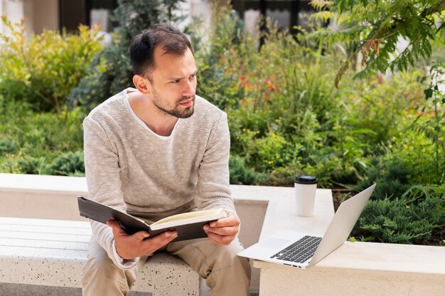 Front view of man working outdoors with laptop and book