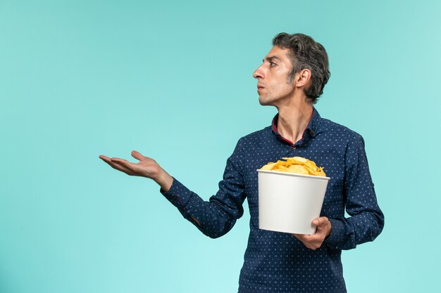 Мужчина средних лет, вид спереди, держит корзину с картофельными чипсами на синем столе