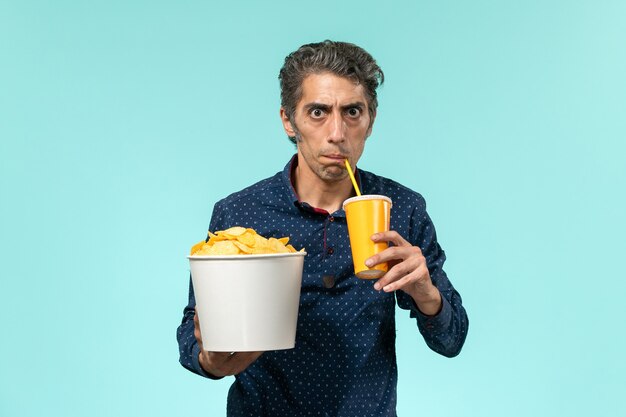 Мужчина средних лет, вид спереди, держащий картофельные чипсы и питьевую соду на синей поверхности
