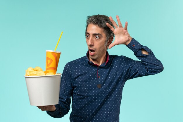 Front view middle-aged male holding potato cips and soda on blue surface