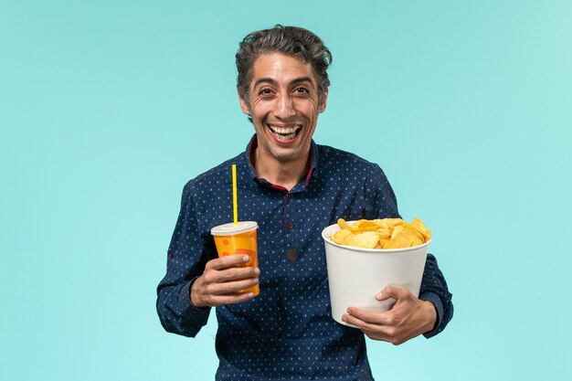 Вид спереди мужчина средних лет, держащий картофельные чипсы и газировку, смеющийся на синей поверхности