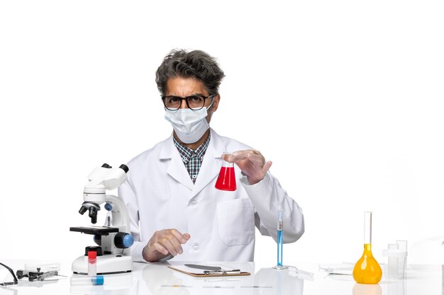 Front view middle-aged scientist in special suit sitting holding flask with red solution