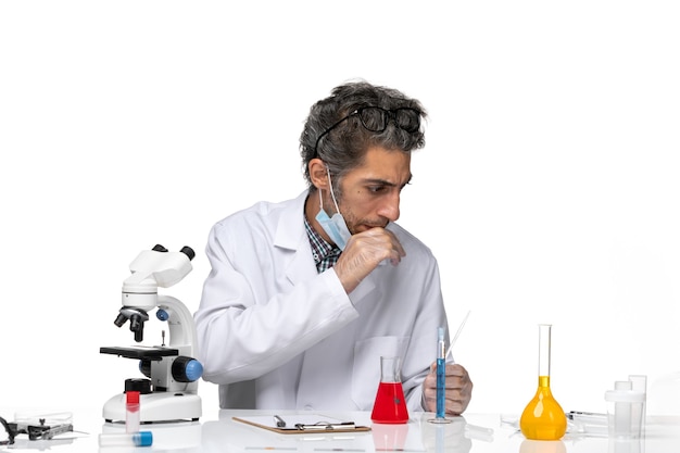 Вид спереди ученого средних лет в белом медицинском костюме, сидящего за столом с растворами