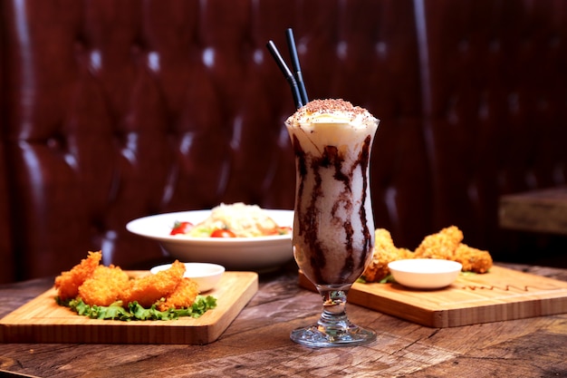 Front view milkshake with whipped cream and chocolate icing with appetizers and salad on the table