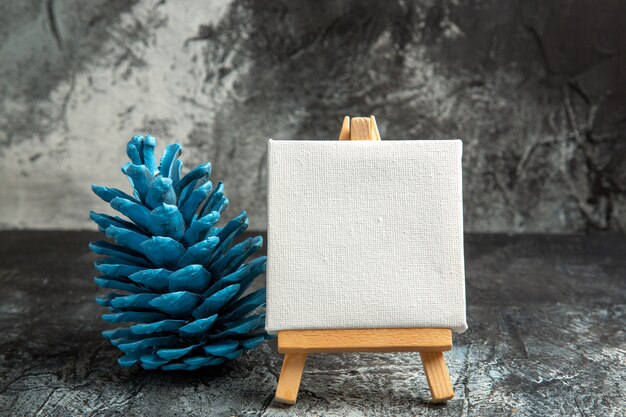 Front view mini white canvas with wooden easel blue pinecone on dark
