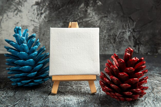 Front view mini white canvas with wooden easel colorful pinecones on dark isolated background