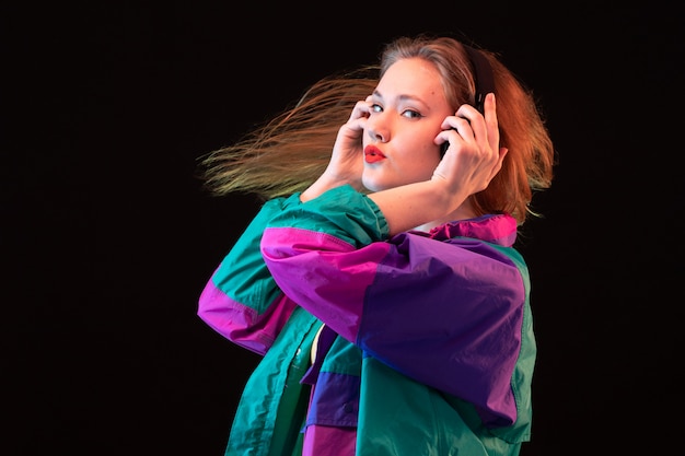 A front view modern young lady in colorful coat orange t-shirt with black earphones posing listening to music on the black background dance modern fashion