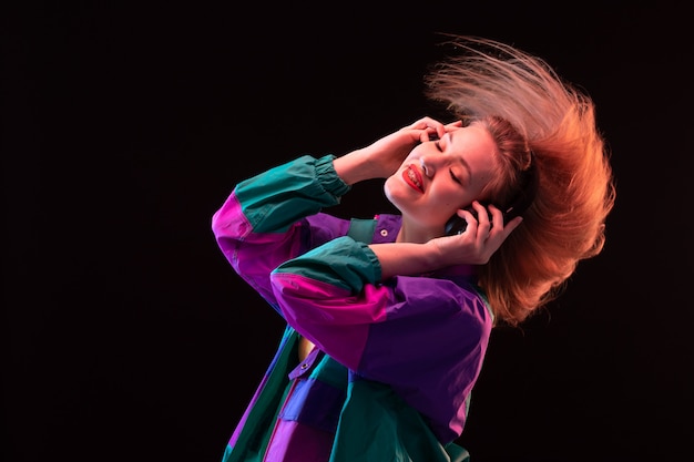 A front view modern young lady in colorful coat orange t-shirt with black earphones posing listening to music on the black background dance modern fashion