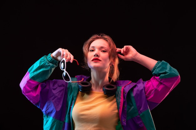 A front view modern young lady in colorful coat orange t-shirt with black earphones and sunglasses posing on the black background dance modern fashion