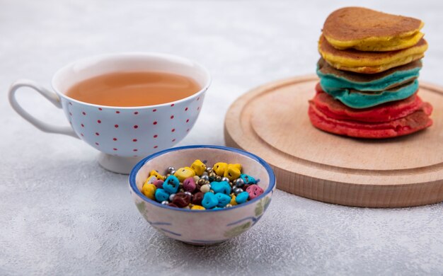 Front view multicolored pancakes on a stand with colored chocolates and a cup of tea on a white background