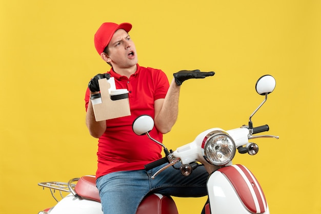 Front view of nervous courier man wearing red blouse and hat gloves in medical mask delivering order sitting on scooter holding orders pointing something on the left side
