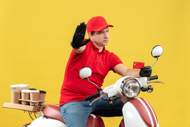 銀行カードを示すスクーターに座って注文を配信する医療マスクで赤いブラウスと帽子の手袋を身に着けている神経質な宅配便の男の正面図
