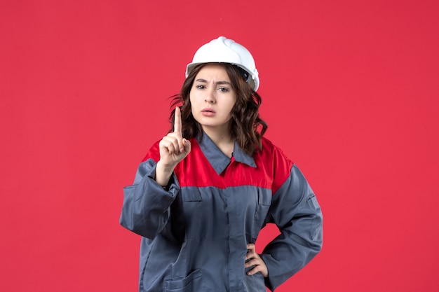 Vista frontale del costruttore femminile nervoso in uniforme con elmetto e rivolto verso l'alto su sfondo rosso isolato
