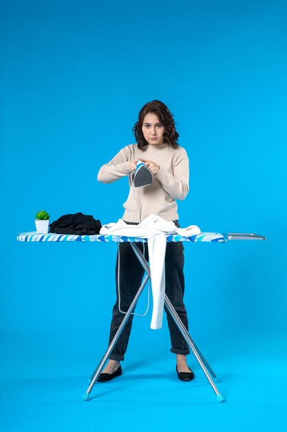 Front view of nervous young female standing behind the board and ironing the clothes on blue background