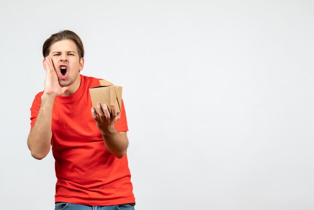 Front view of nervous young guy in red blouse holding small box calling someone on white background