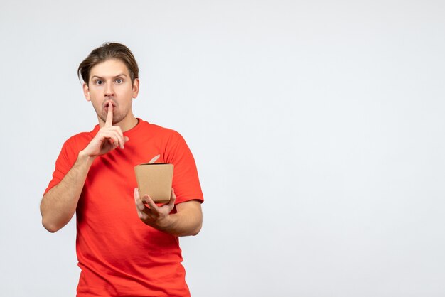 Вид спереди нервного молодого парня в красной блузке, держащего маленькую коробку, делая жест молчания на белом фоне
