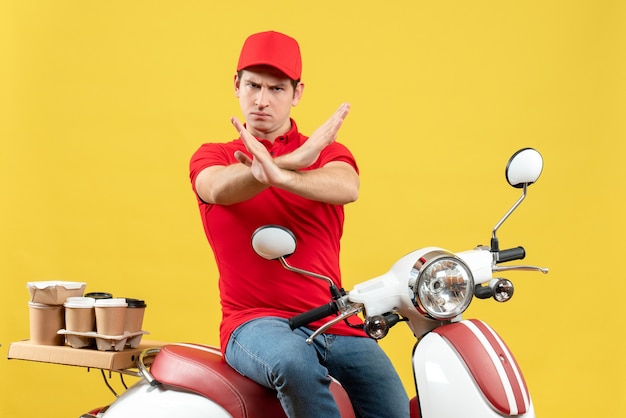 Вид спереди нервного молодого парня в красной блузке и шляпе, доставляющего заказы, делая жест стоп на желтом фоне