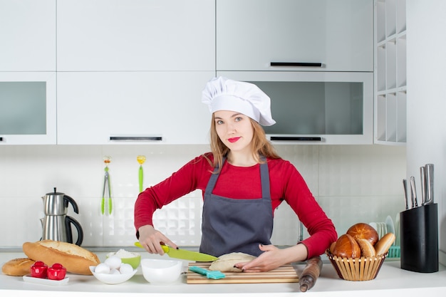 Front view nice woman in cook hat and apron cutting bread in modern kitchen