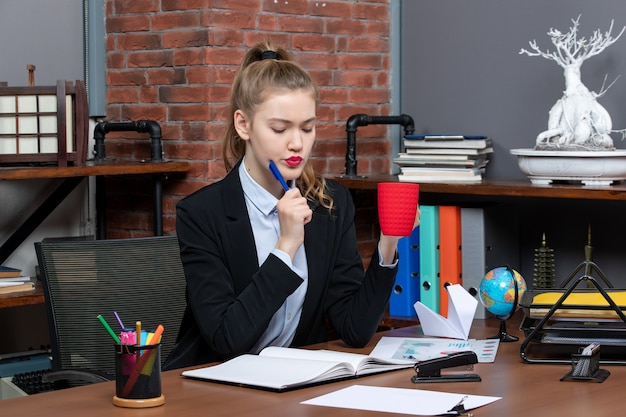 テーブルに座って、オフィスで彼女の前に赤いカップ読書文書を保持している忙しい若い女性の正面図