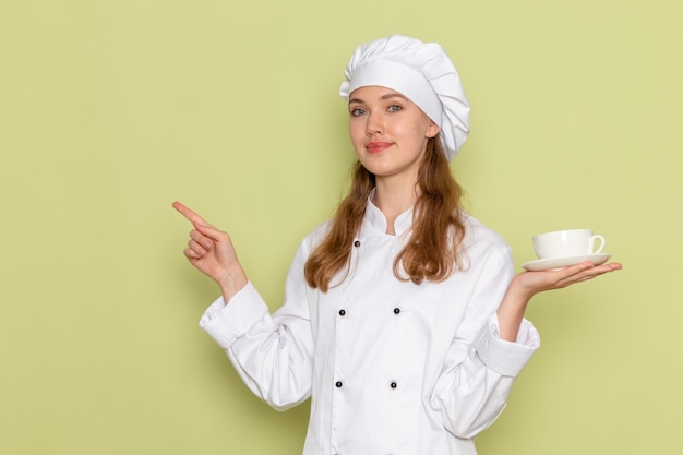 Бесплатное фото Вид спереди повара в белом костюме повара, держащего чашку кофе и улыбающегося на зеленом столе, кухня, приготовление еды, женский цвет
