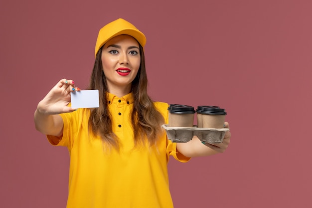 Бесплатное фото Вид спереди курьера в желтой форме и кепке, держащего коричневые кофейные чашки и открытку на розовой стене