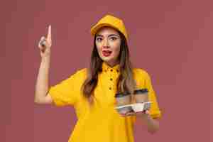 Бесплатное фото Вид спереди курьера-женщины в желтой форме и кепке, держащего кофейные чашки на розовой стене