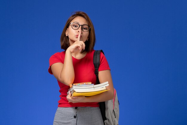 밝은 파란색 벽에 책과 파일을 들고 배낭과 빨간 셔츠에 여성 학생의 전면보기
