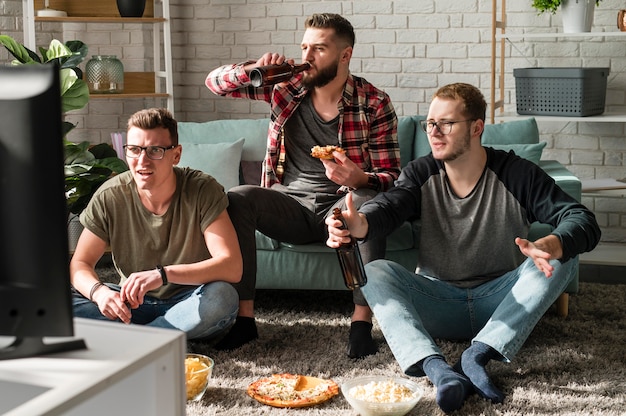 Бесплатное фото Вид спереди друзей-мужчин, которые едят пиццу и смотрят спорт по телевизору с пивом