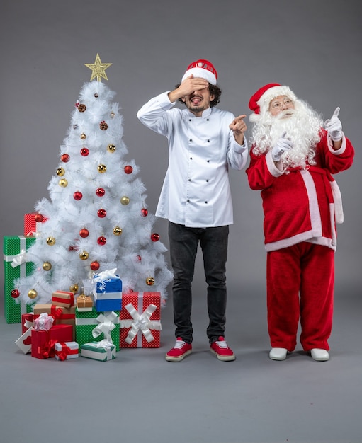 Бесплатное фото Вид спереди санта-клауса с мужчиной-поваром вокруг рождественских подарков на серой стене