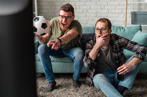 一緒​に​テレビ​で​スポーツ​を​見て​、​サッカー​を​している​2​人​の​陽気な​男性​の​友人​の​正面図