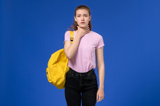 파란색 벽에 목이 아픈 노란색 배낭을 입고 분홍색 티셔츠에 젊은 여성의 전면보기